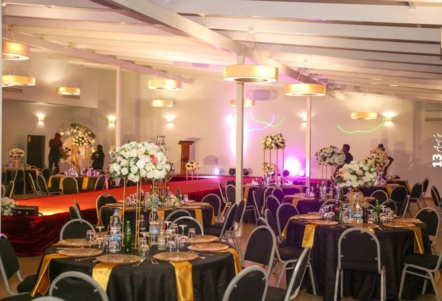 Salle de réception dressée avant l’arrivée des invités : tables rondes, nappage sombre et chemins dorés, tribune éclairée en fond